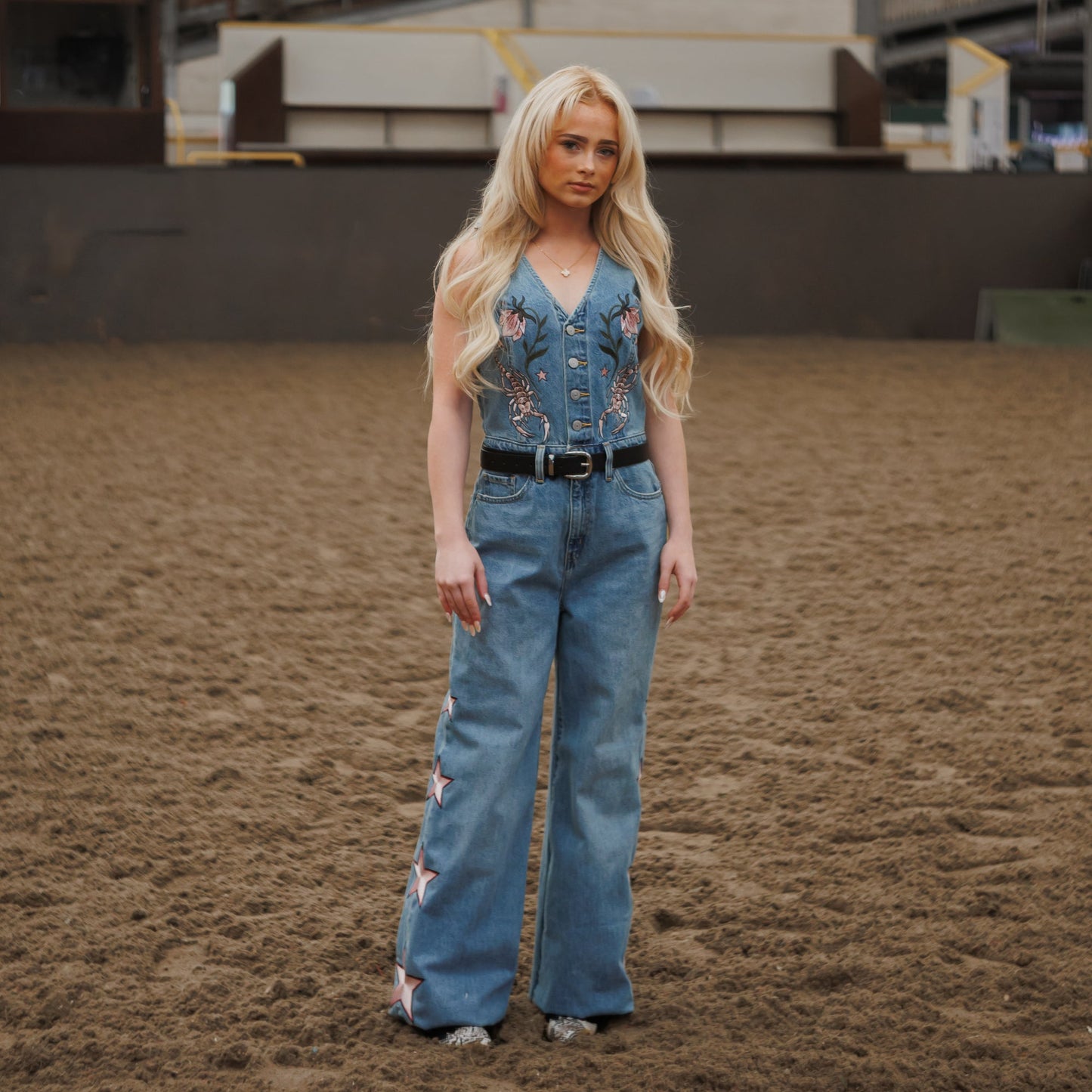 A blonde woman stands in a sandy indoor arena wearing a blue denim vest and matching wide-leg jeans with floral embroidery and a black belt.