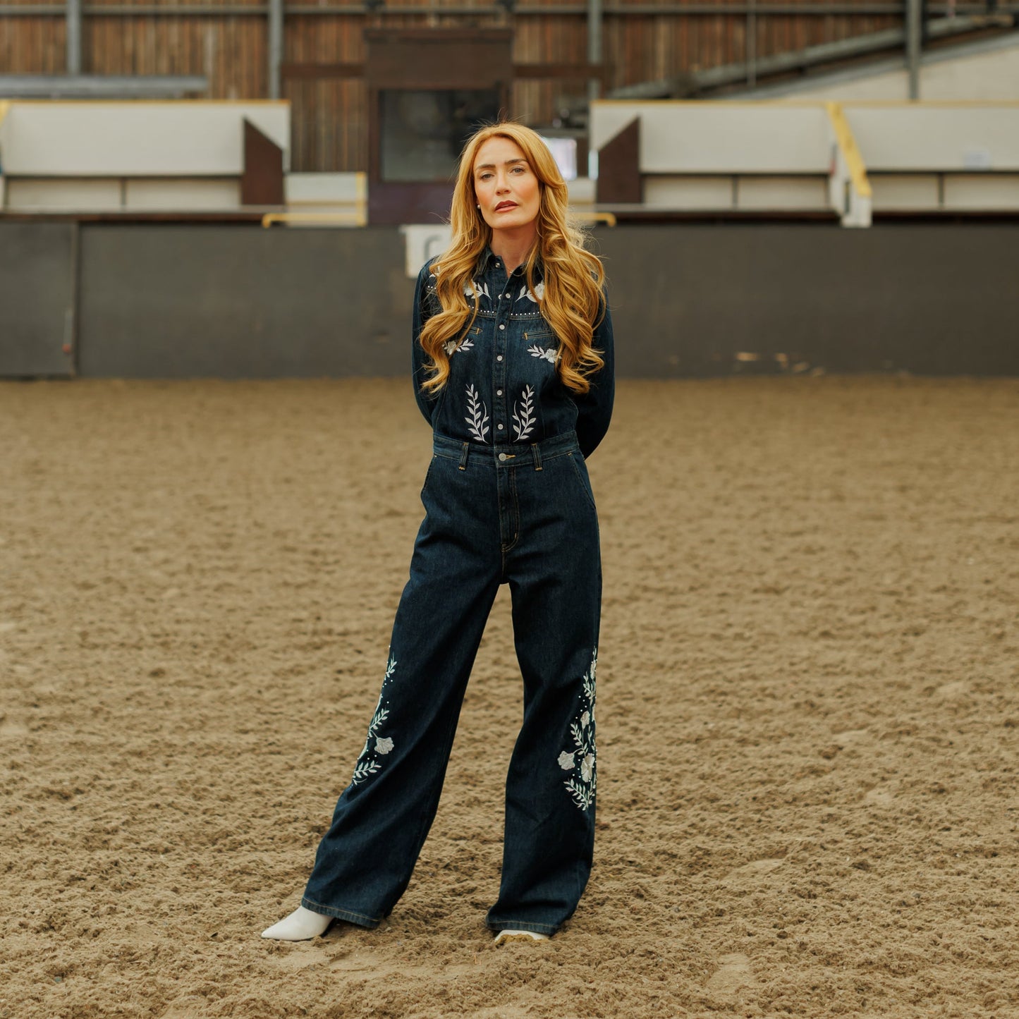 A woman stands in a sandy indoor arena wearing a dark denim western jumpsuit with floral embroidery on the chest and legs, paired with white boots.