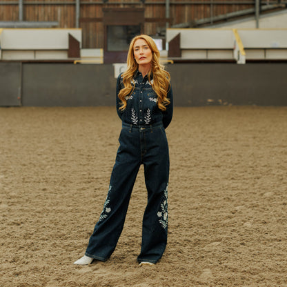 A woman stands in a sandy indoor arena wearing a dark denim western jumpsuit with floral embroidery on the chest and legs, paired with white boots.