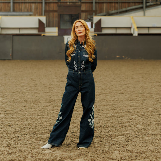 A woman stands in a sandy indoor arena wearing a dark denim western jumpsuit with floral embroidery on the chest and legs, paired with white boots.