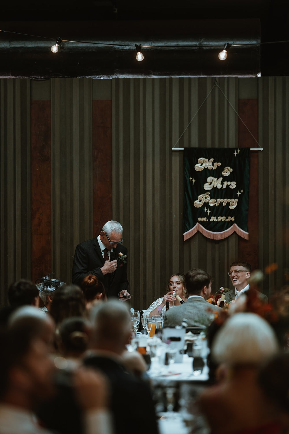 A wedding reception scene with people laughing. A luxury embroidered wall hanging saying "Mr & Mrs Perry" + wedding date is hanging on the main wall. 