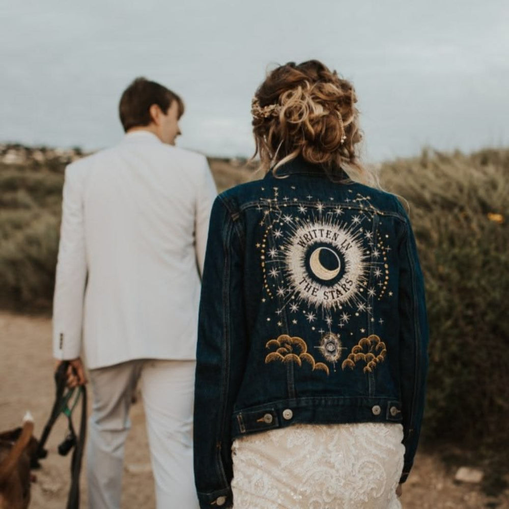 A woman seen from behind wearing a dark blue denim jacket with a large celestial moon and star embroidered on the back, walking outdoors with a man in a white suit.