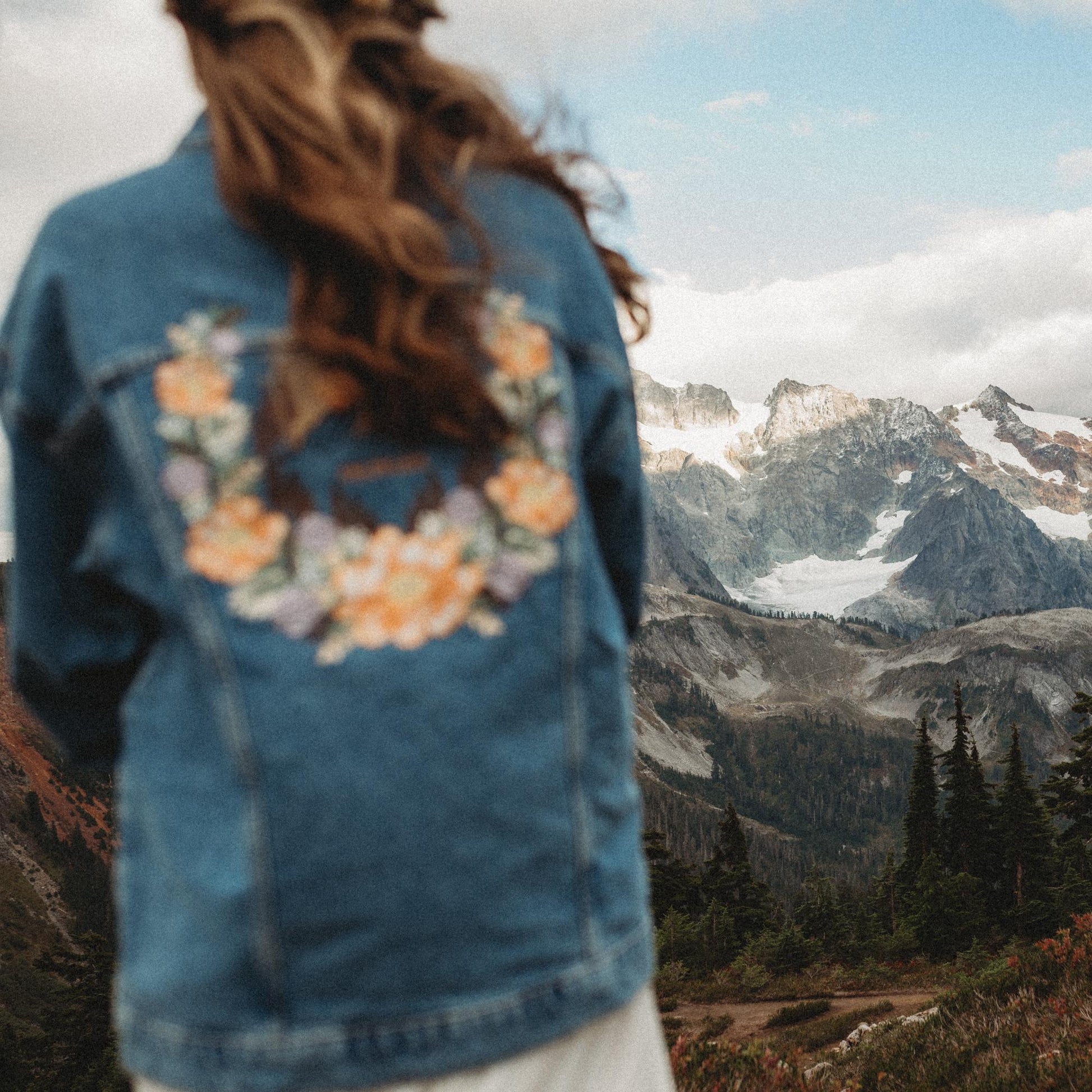 Person wearing a denim jacket with floral embroidery, standing in front of a mountain landscape.