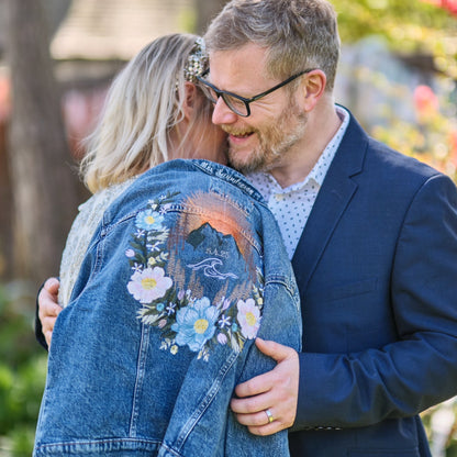 Bride and groom embracing. Bride is holding a blue denim jean jacket on her shoulder with the embroidery design of a mountain floral wreath and wave feature stitched on the back. 