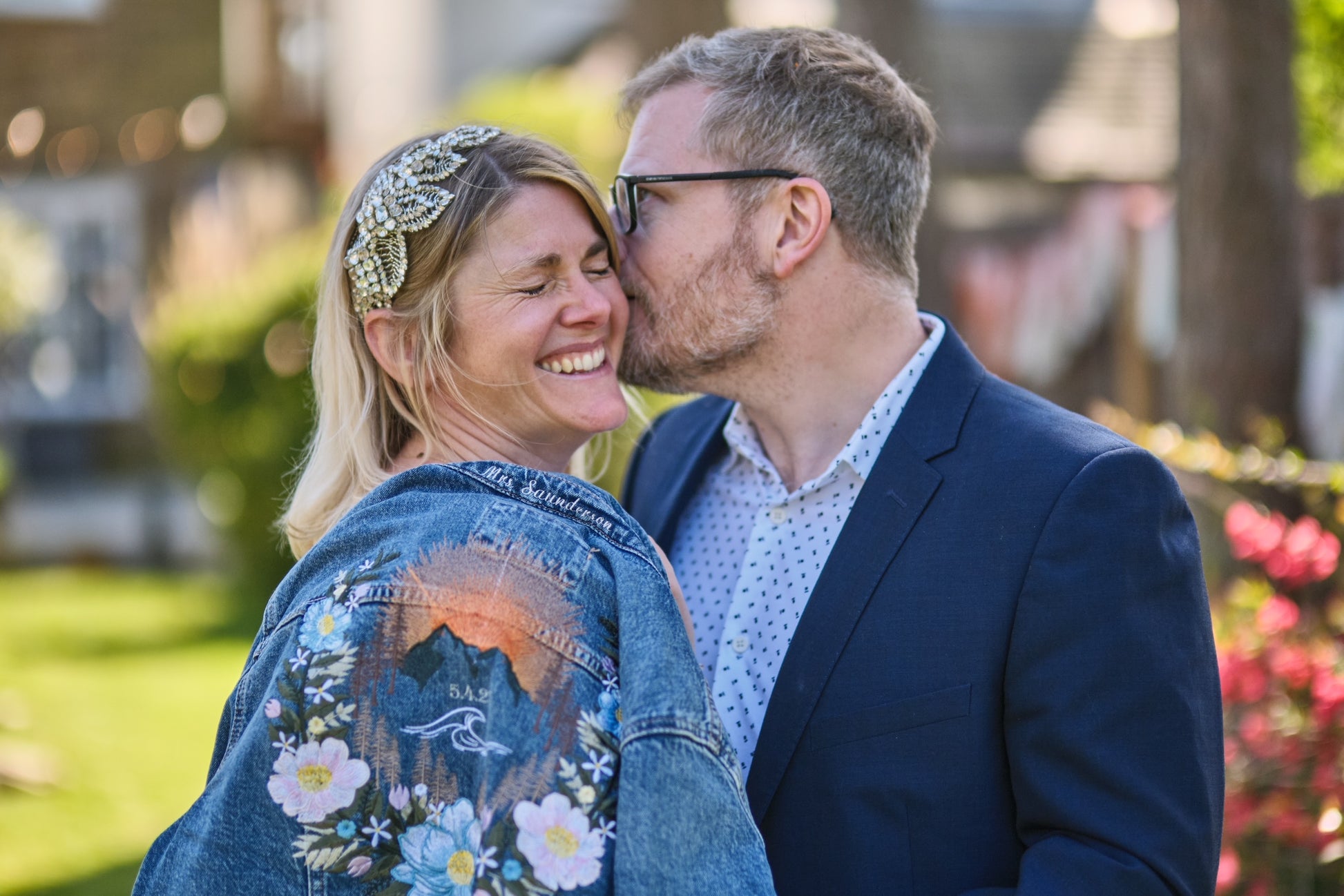 A smiling blonde woman wearing a decorative gold headband and a blue denim jacket with a mountain and floral print, being kissed on the cheek by a man in a navy blazer and light blue patterned shirt, set against a blurred outdoor garden background.