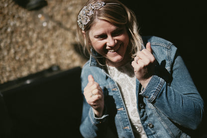 A smiling woman wearing a sparkly silver headband and a blue denim jacket over a white top, giving a thumbs-up in a low-angle outdoor shot.