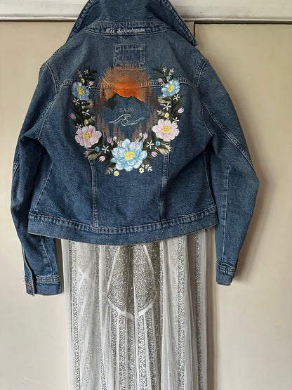 A brides outfit hung up on a door - featuring a sparkly wedding dress with denim jacket over the top. The jacket is light blue denim and has a floral mountain design which is embroidered.
