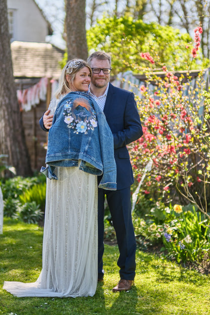 A portrait photo of a bride and groom. The bride is wearing a white sparkly dress with a light blue denim jacket - a floral mountain floral design customised and embroidered by Beth Wilson Embroidery.