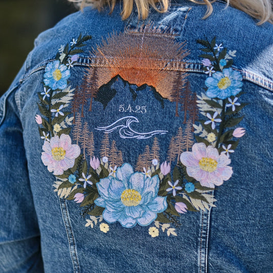 Close-up of the back of a blue denim jacket worn by a person, featuring a large embroidered floral wreath with pink, blue, and white flowers around a mountain and wave design with the text 'E.A.
