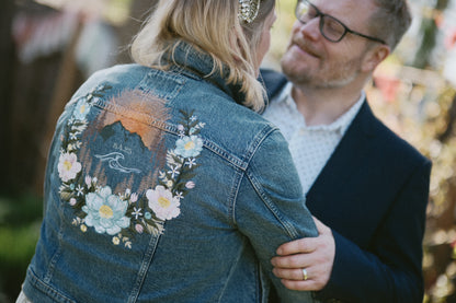 A close up of a custom floral mountain jacket embroidered by Beth Wilson Embroidery on a medium blue jean denim jacket. The design features a mountain, sunrise and wave motif with pastel florals 